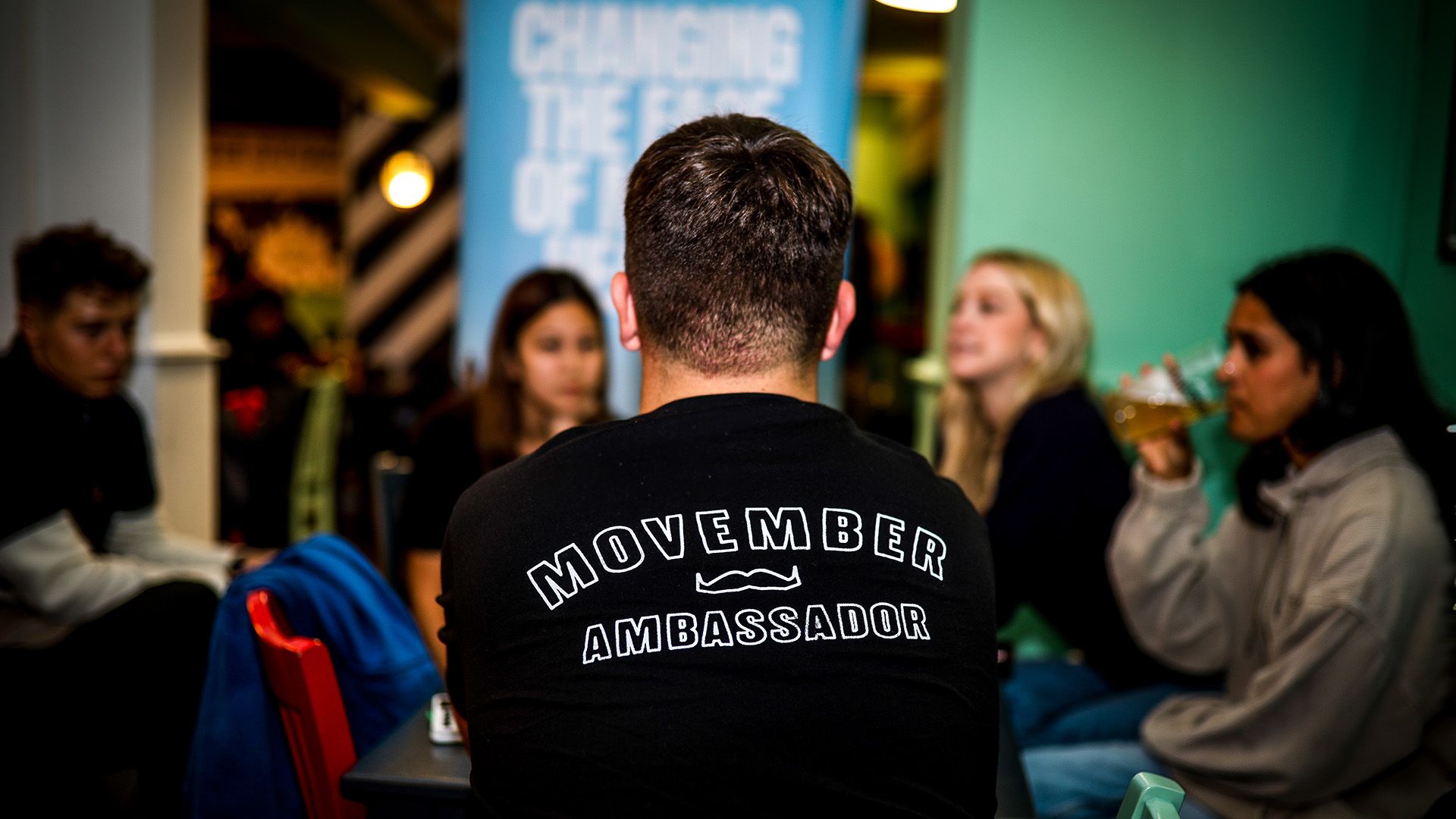 Photo of a man at a party, wearing a black shirt that says: "Movember ambassador".