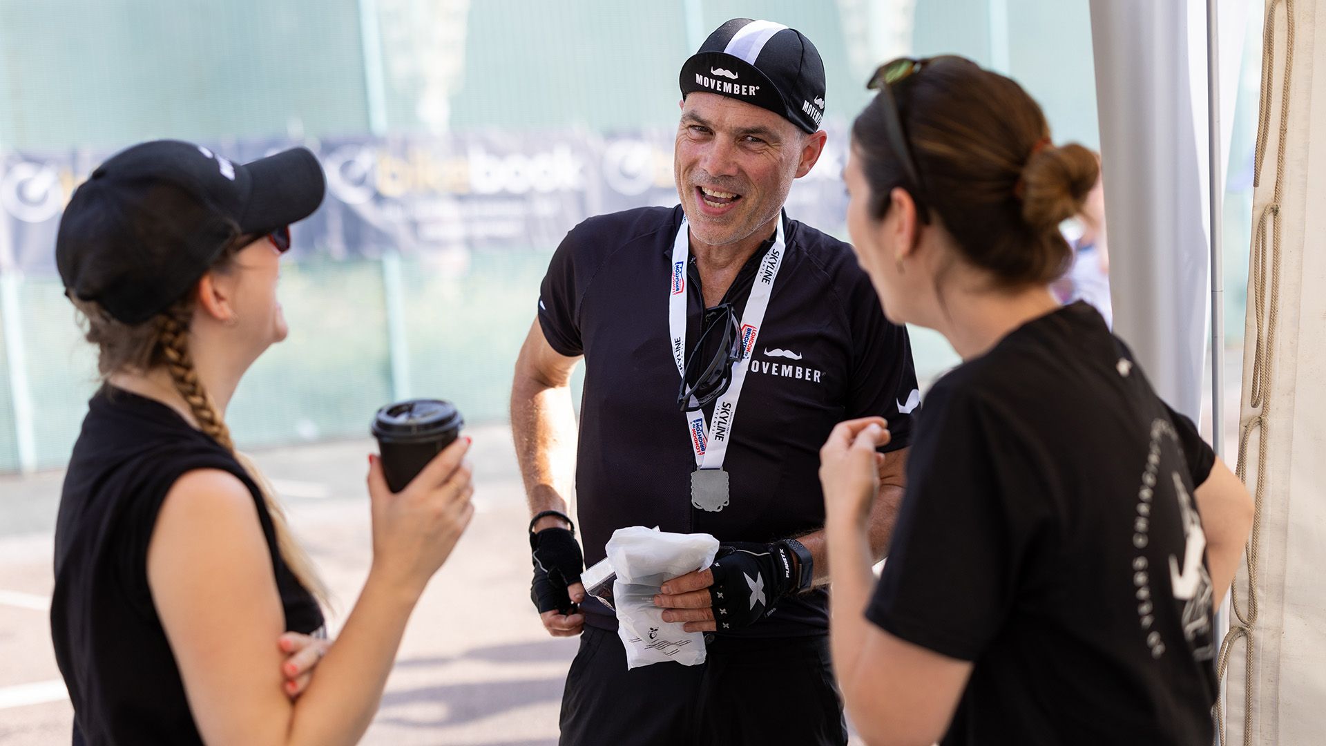 Photo of smiling cyclist after a race chatting to two people wearing black Movember-branded attire.