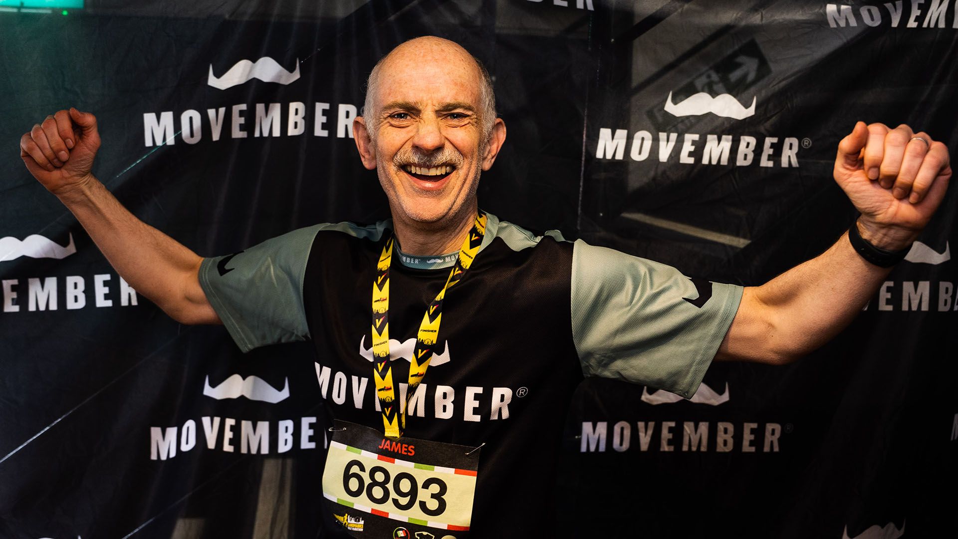 Photo of runner in Movember-branded running gear, triumphantly lifting his arms to camera after finishing a race.