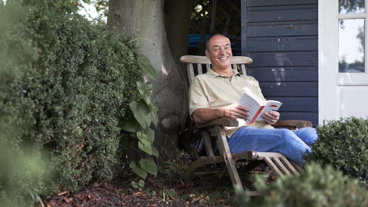 Photo of smiling man on his porch, looking up to camera from reading his book.