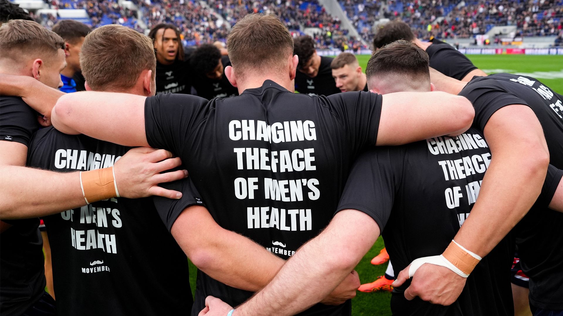 A group of men with arms around eachother and text on shirts that say "Changing the Face of Men's Health"