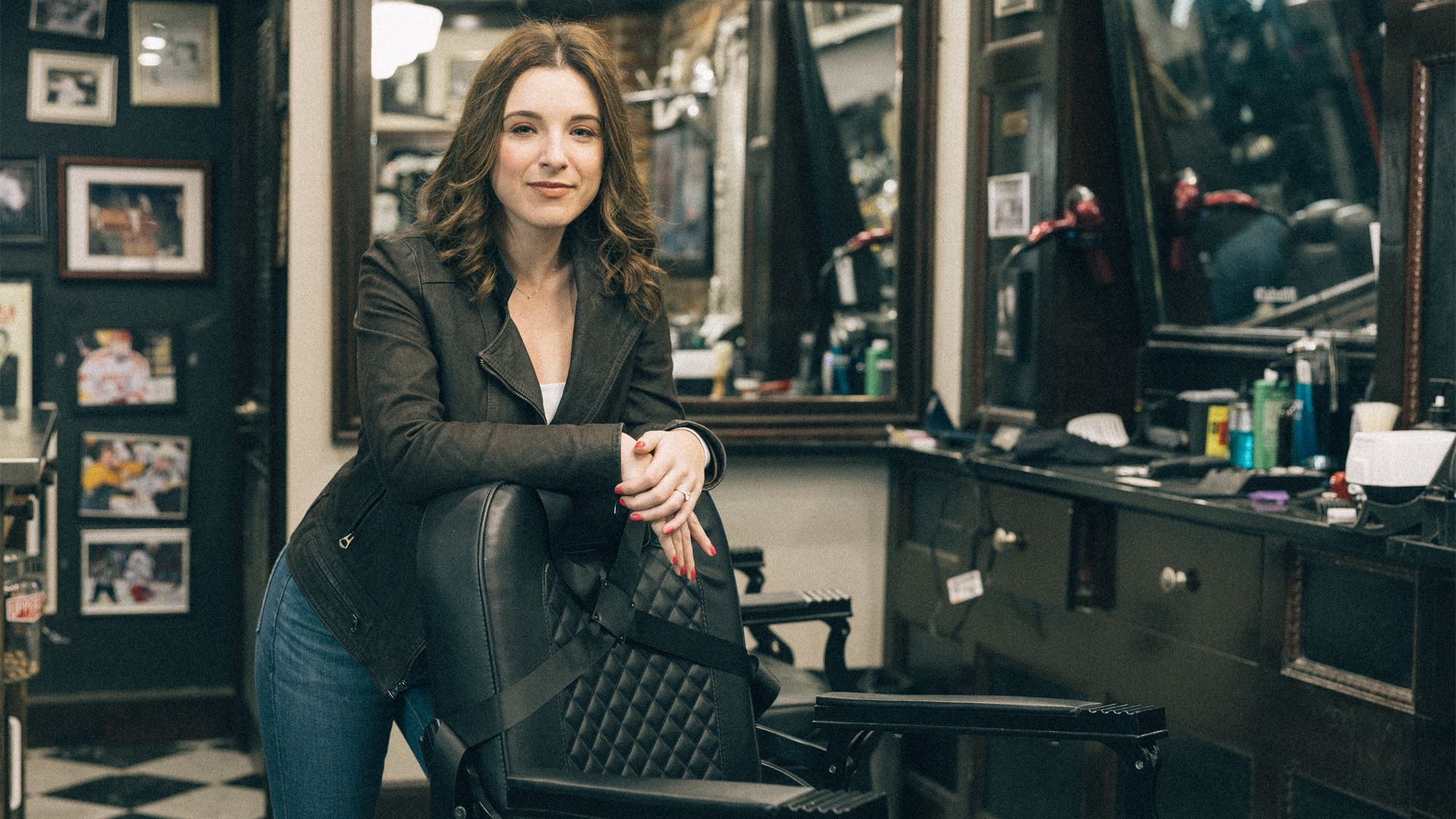 A woman poses for a portrait standing behind an empty barber chair in a barber shop