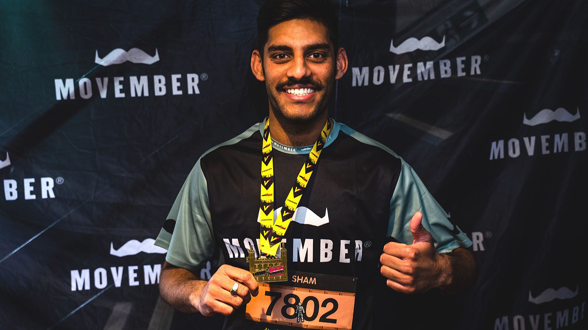 Photo of marathon runner in Movember-branded running gear, triumphantly giving a thumbs up to camera after finishing a race..