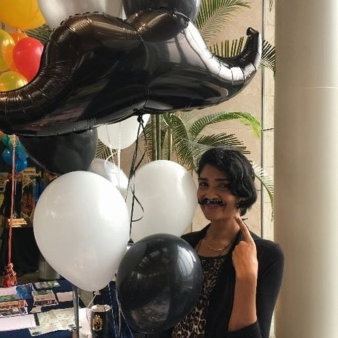 Photo of a woman next to a moustache-shaped balloon.