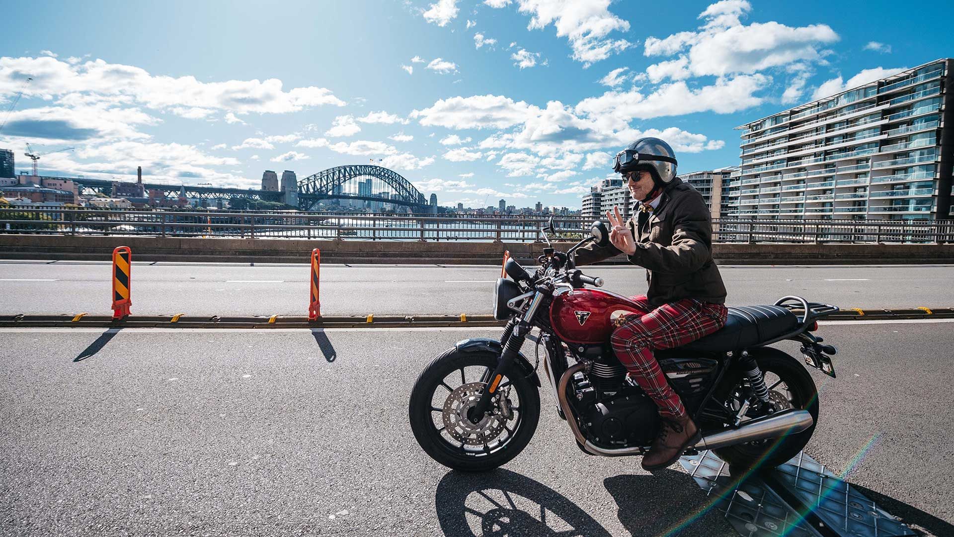 Motorcyclist dressed in dapper attire, saluting the camera with a V-sign. The Sydney Harbour Bridge is in the background.