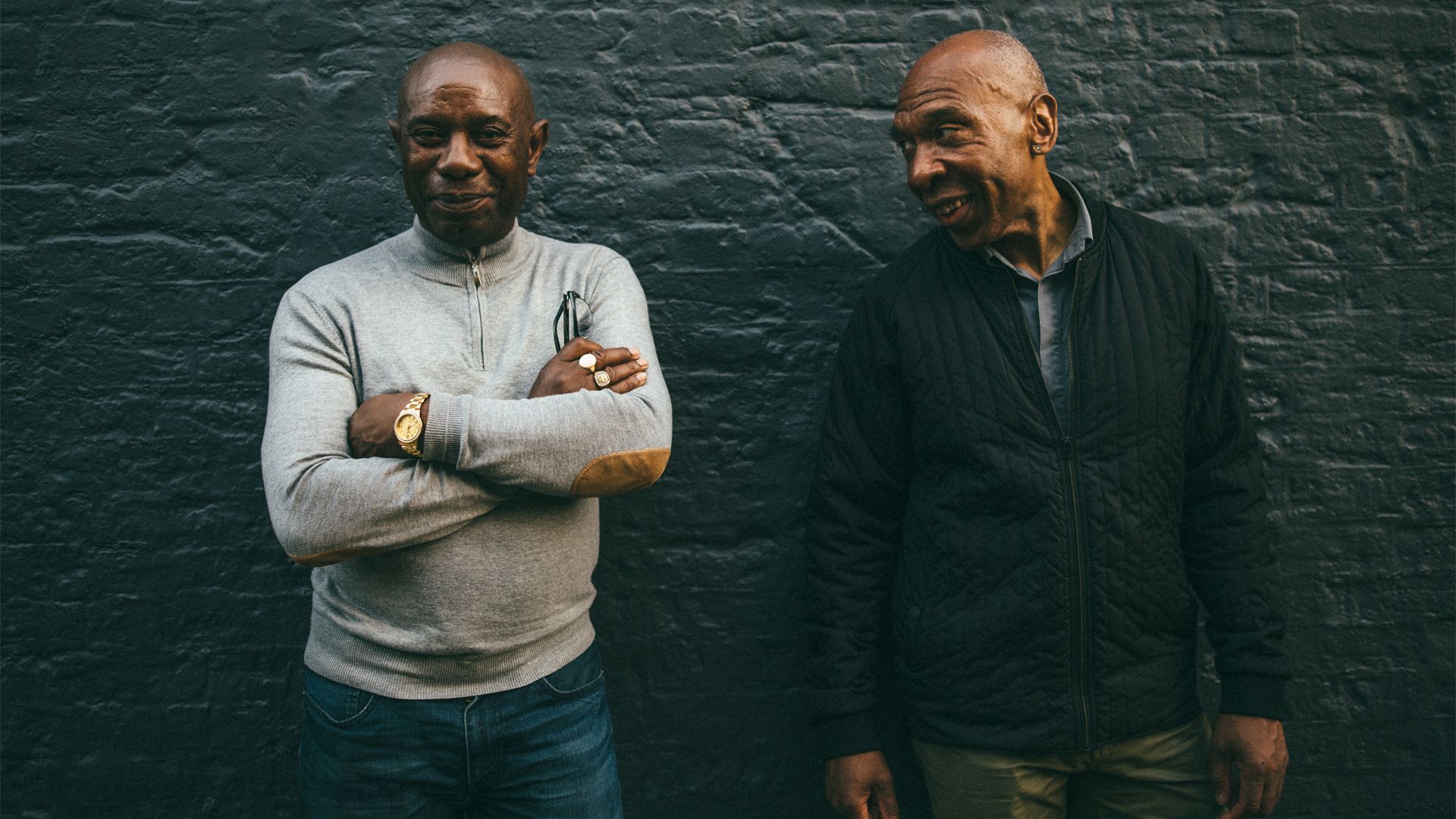 Two men, one with his arms crossed, pose in from of a brick wall