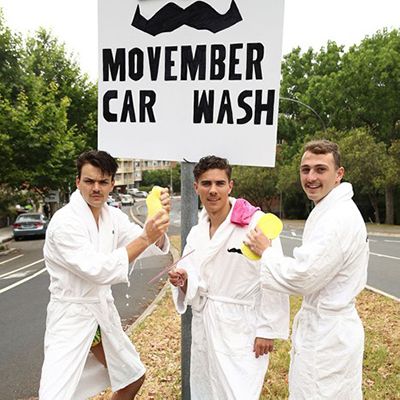 Photo of three young men on a street in dressing gowns. They carry sponges and stand behind a sign that says "Movember car wash".