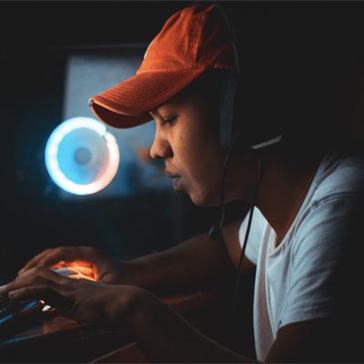 A young person with headphones on sitting in a dark room, lit by the screen of their computer.