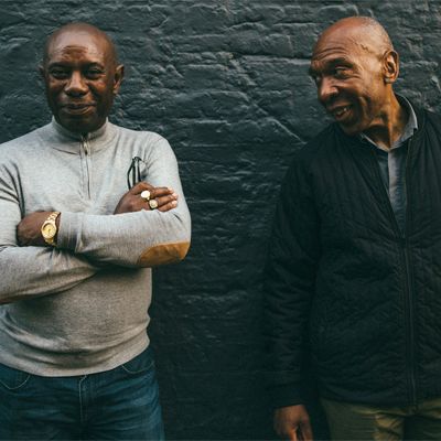 A photo of two senior men standing against a brick wall with slight smiles.