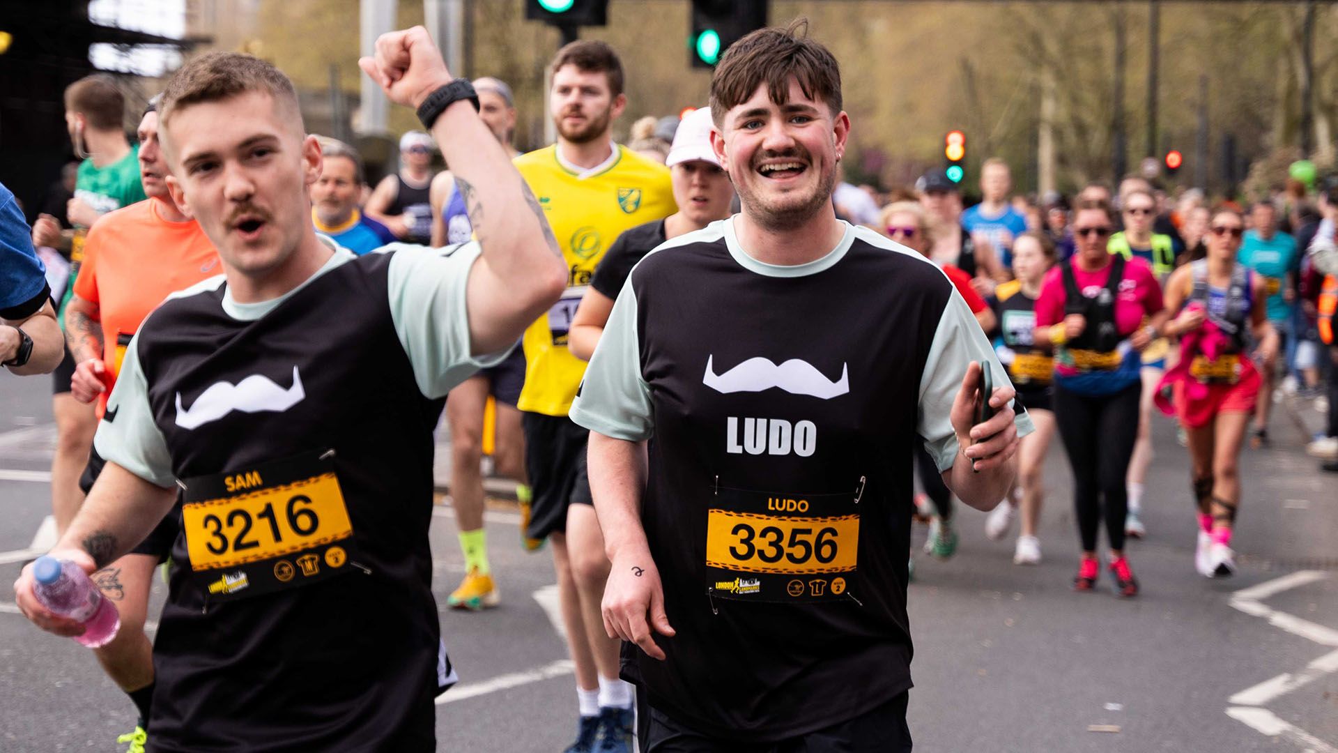 Photo of two young men running a public race, looking triumphantly to camera and wearing Movember-branded running gear.
