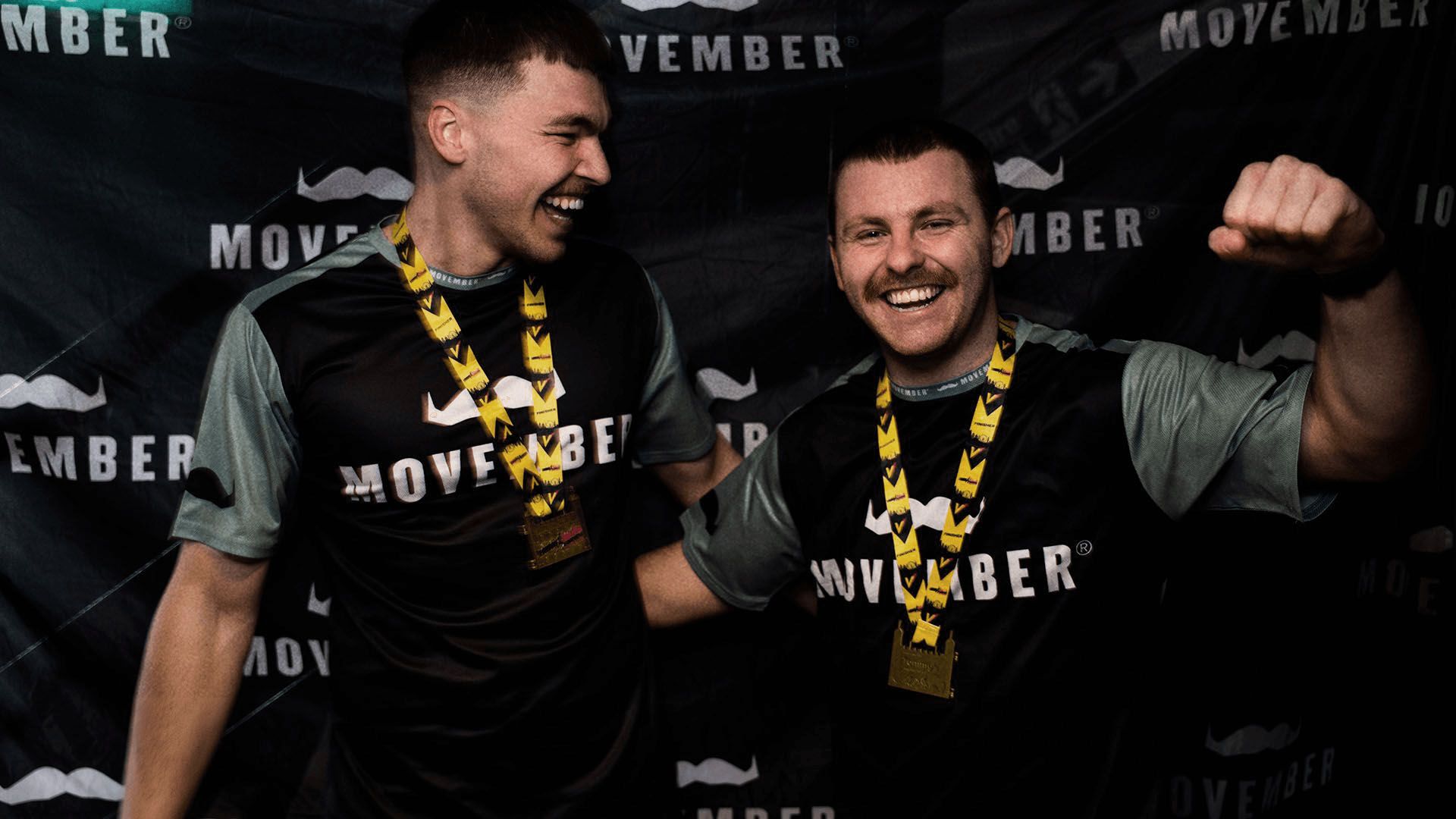 Two marathon runners in Movember-branded attire, triumphantly posing in front of Movember banner after finishing their race.