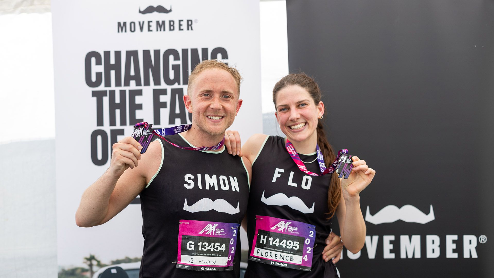 Photo of two runners holding their medals in front of a Movember sign.