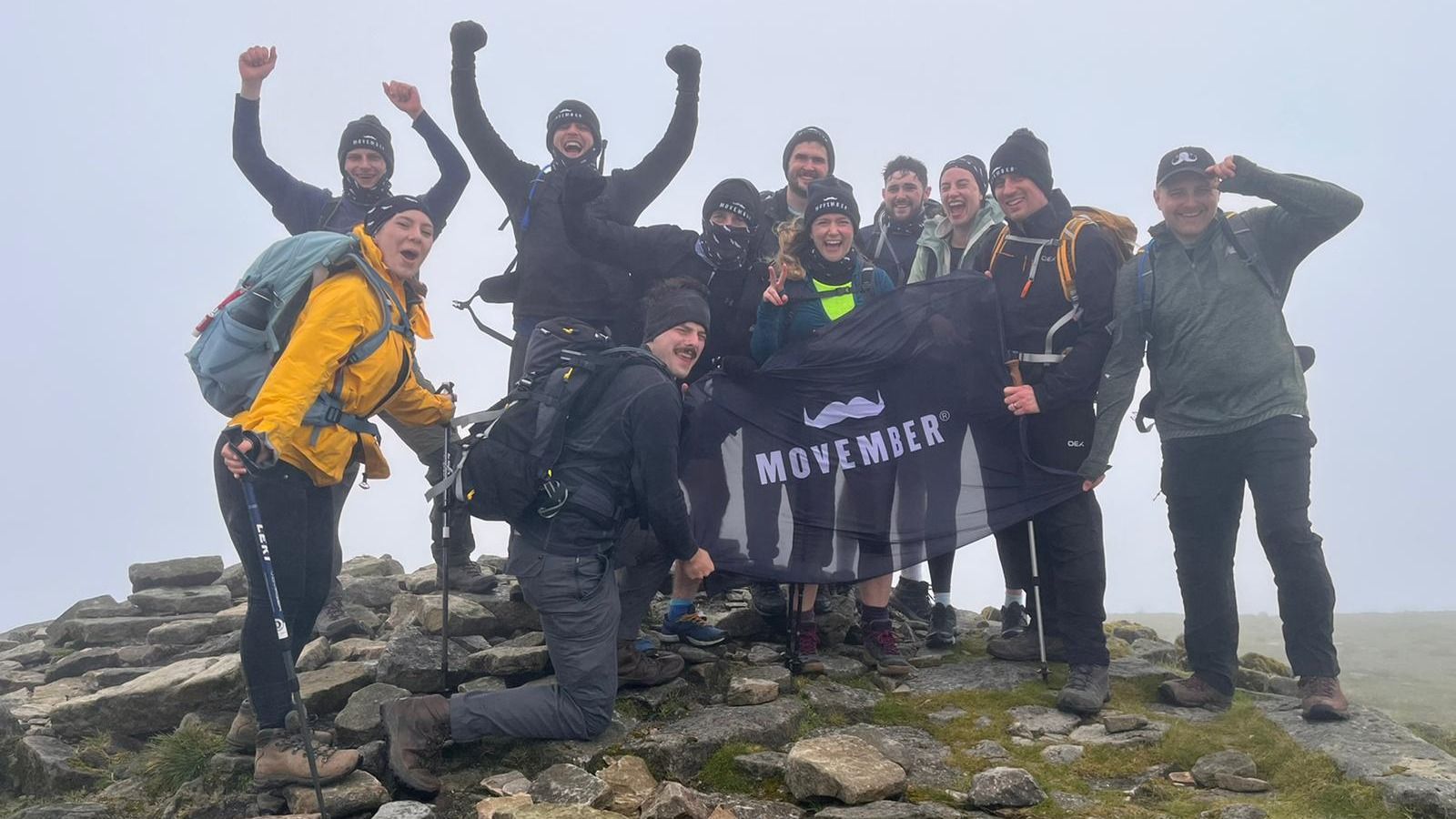 Group photo of the team that took part in the challenge, stood at the top of a peak with a Movember flag