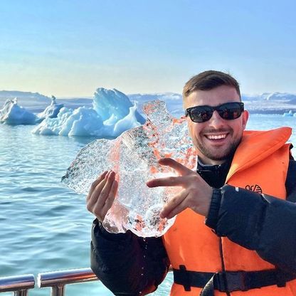 Photo of a smiling man posing before of an arctic ocean landscape, while holding up a block of sea ice.