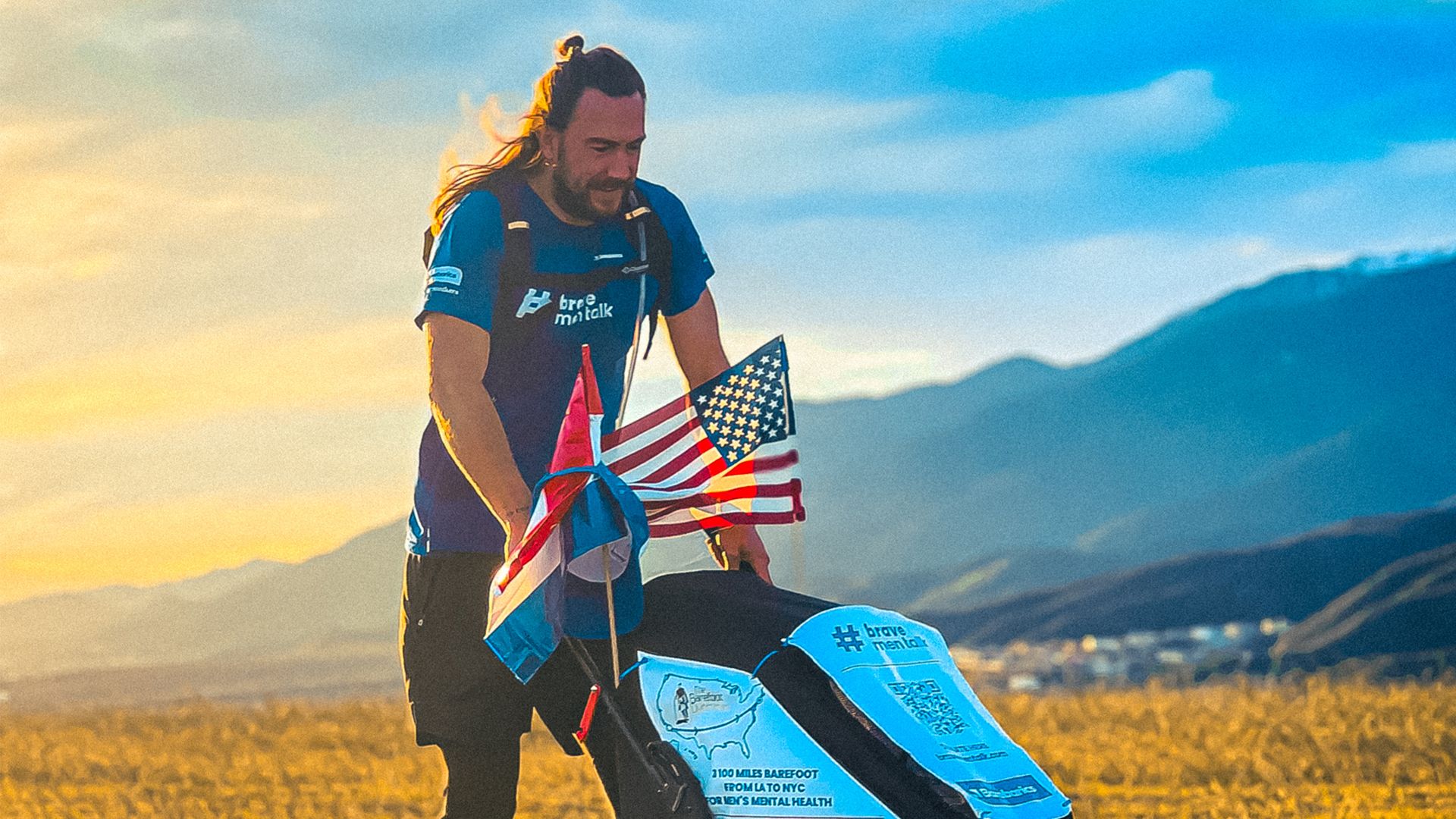 Photo of a man in activewear, pushing a pram clad in American flags. A hazy mountain landscape is visible behind him.