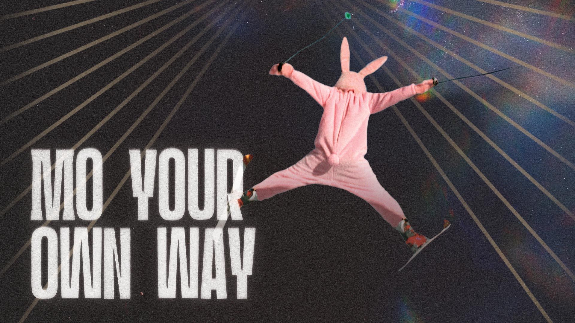 A black and white photo of a man jumping in skies while wearing a full rabbit suit. The words "MO YOUR OWN WAY" are superimposed.