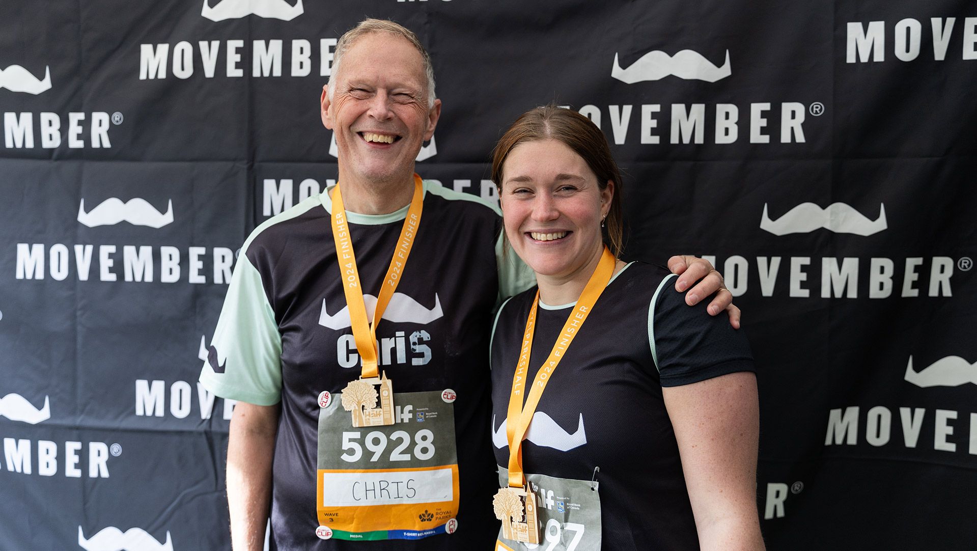 Photo of two runners posing for a photo after a marathon in front of a Movember sign.