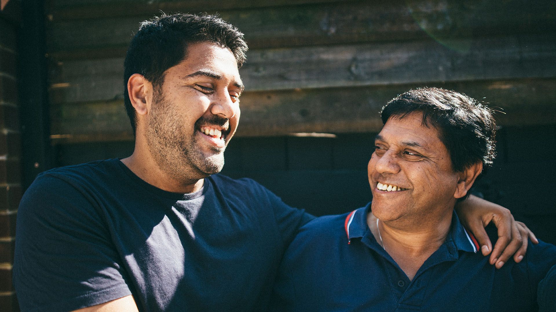 Photo of two smiling middle-aged men looking at each other. One has his arms over the shoulder of the other.