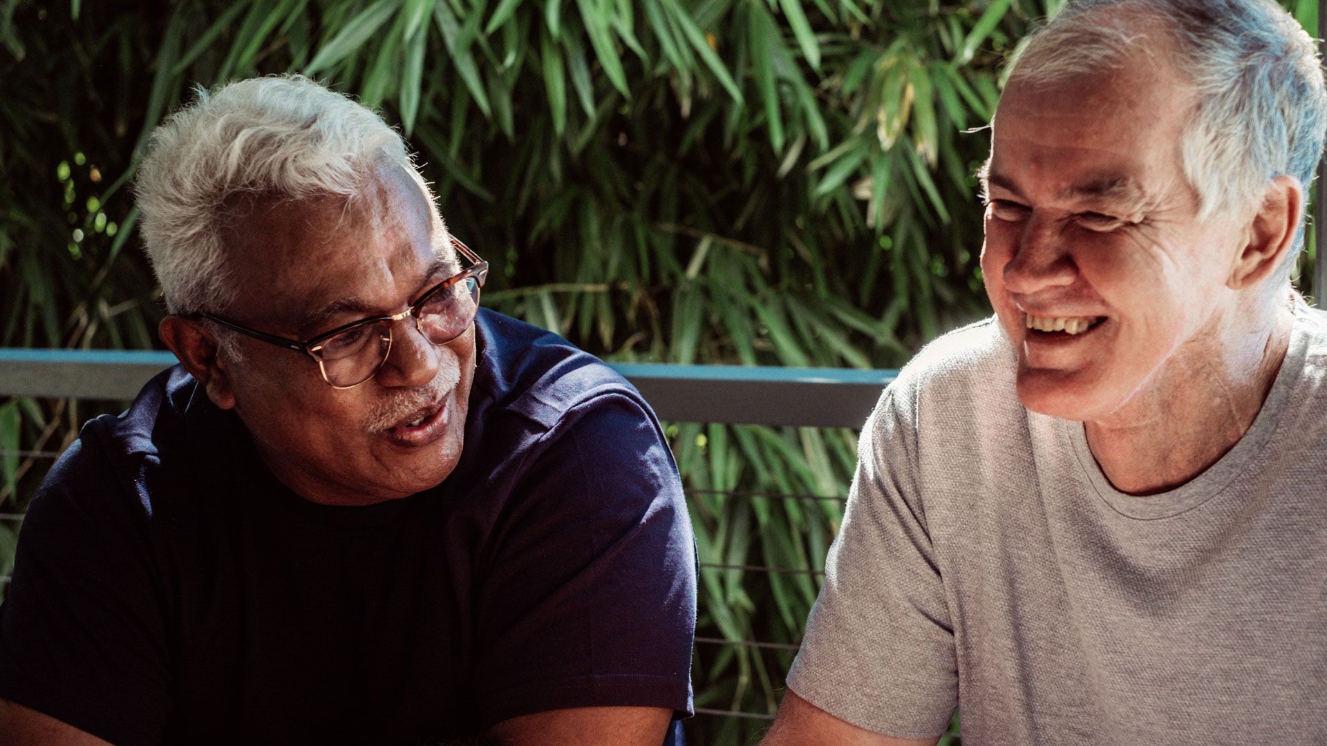 Two older men with moustaches one wearing glasses, sit outside next to each other, both smiling and laughing