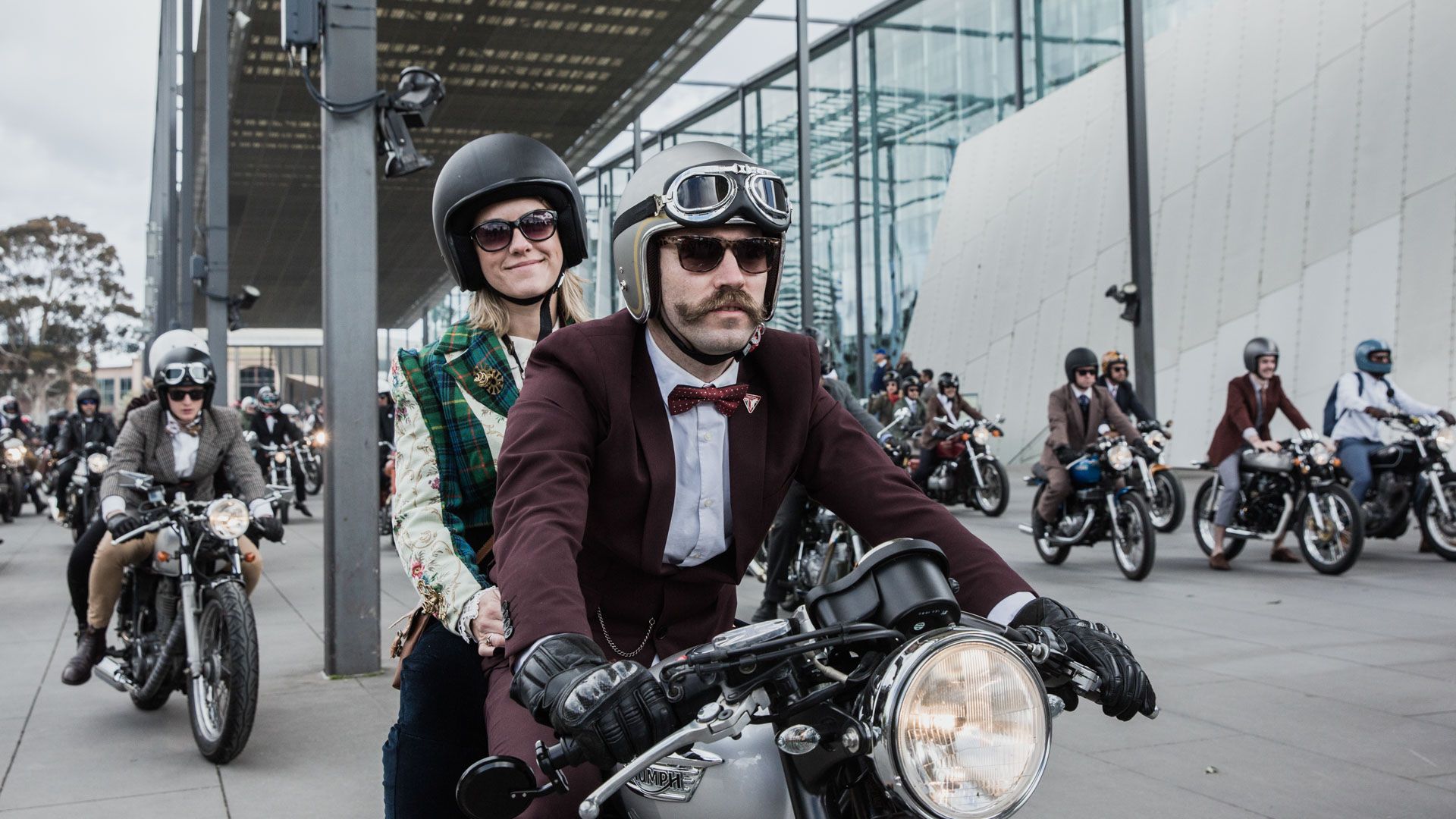 Man with moustache, dressed in suit, sits on motorbike, with female behind on the back of bike. Other motorbike riders stand around them on their bikes. 