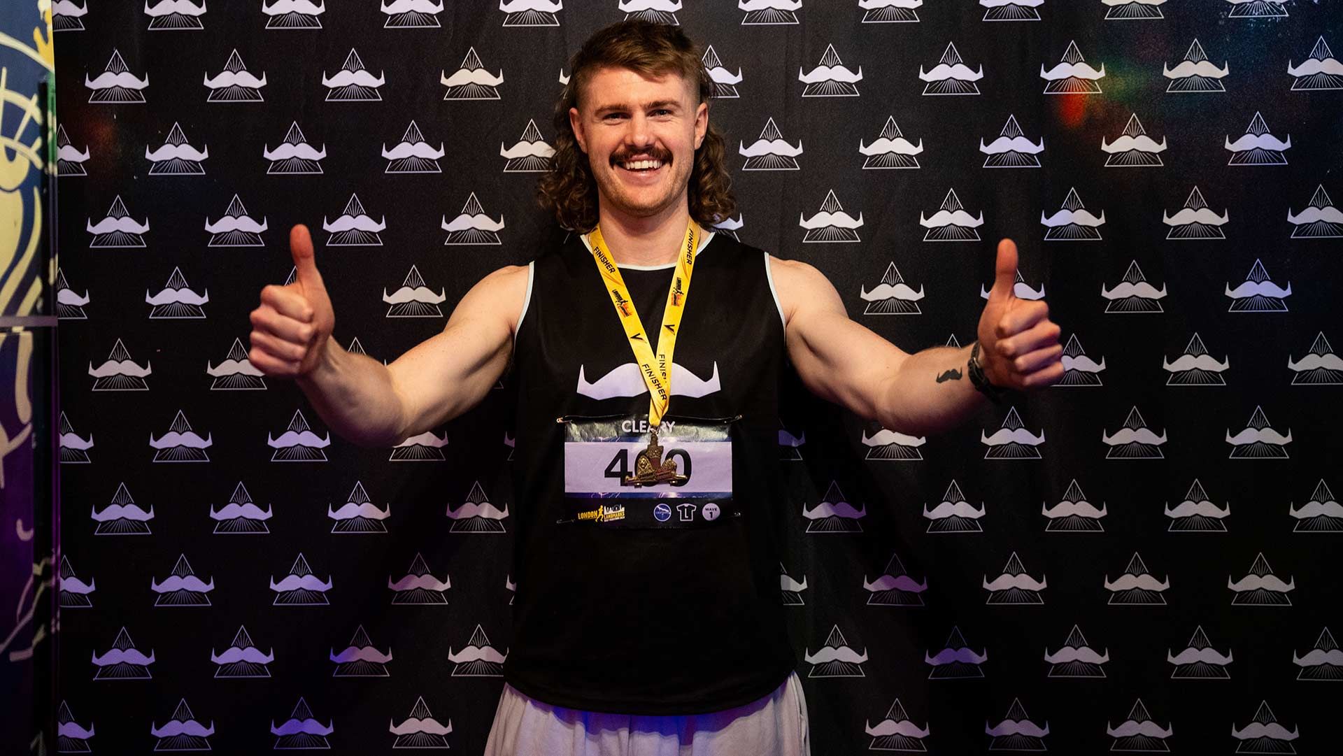 Photo of an athletic young marathon runner triumphantly throwing thumbs up to camera in front of Movember signage.